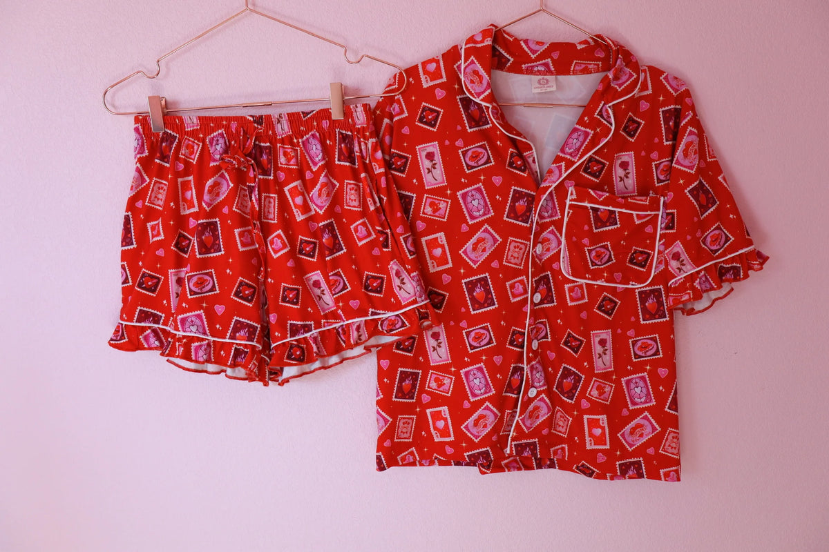 A two-piece pajama set consisting of a short-sleeve button-up shirt and matching shorts, both in a vibrant red fabric with a playful pattern of pink, purple, and black playing cards, hanging on a light pink wall.