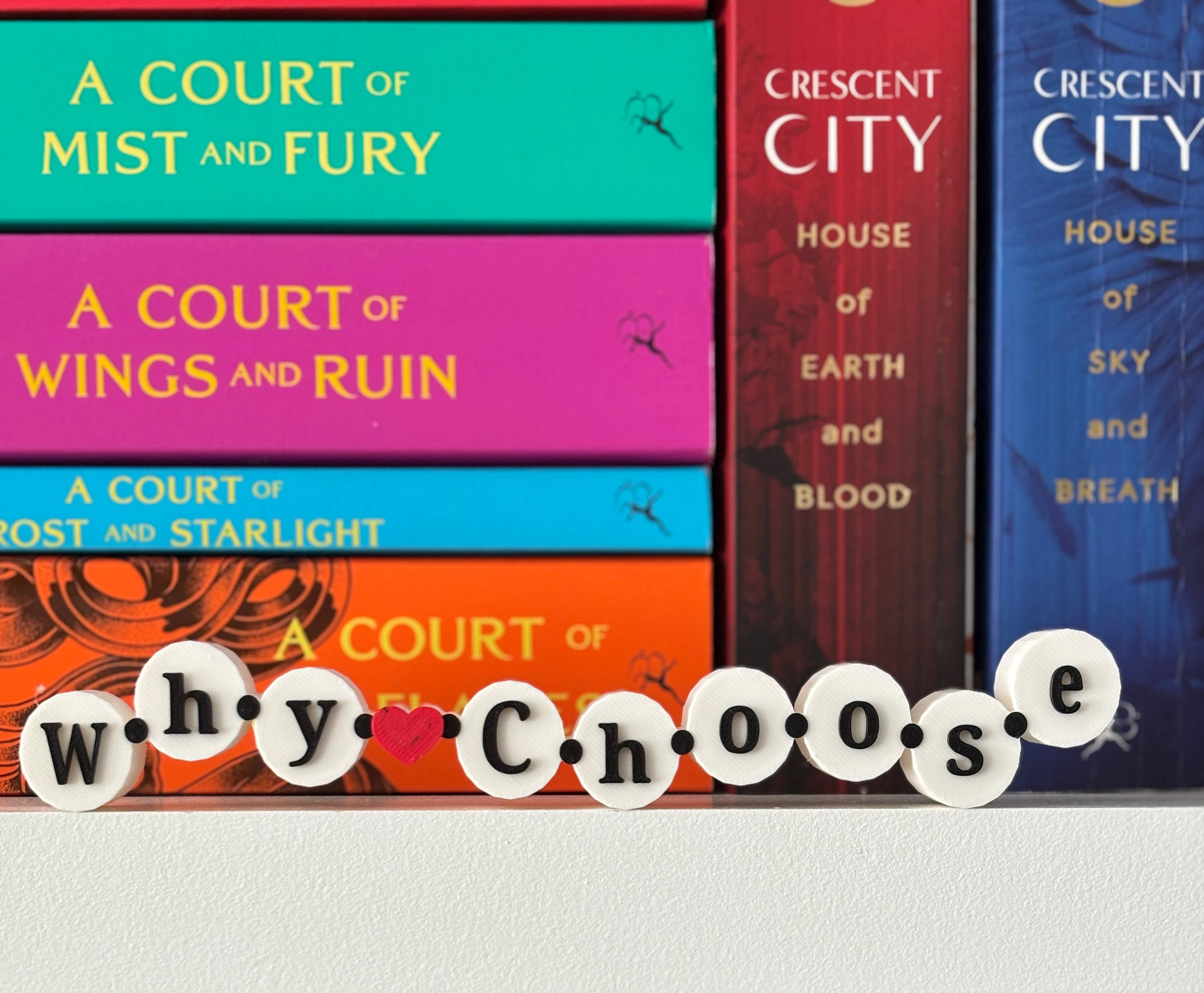 A stack of colorful romance novel book spines with decorative white letter tiles spelling 'why choose' placed in front on a light surface.